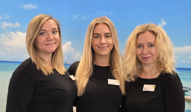 Drei lächelnde Frauen mit blonden Haaren stehen vor einem Strandhintergrund