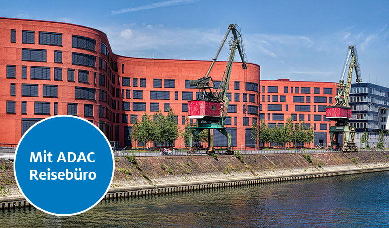 Rotes Gebäude mit Kränen am Flussufer, klarem Himmel und Bäumen.