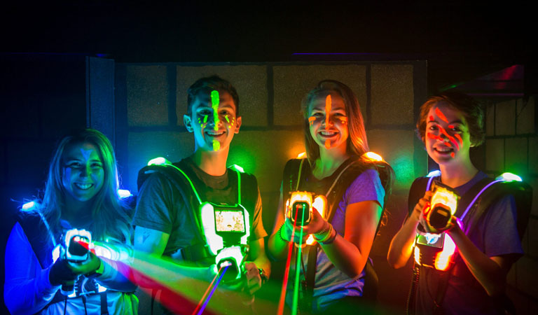 Vier junge Menschen mit leuchtenden Westen lächeln beim Lasertag im dunklen Raum