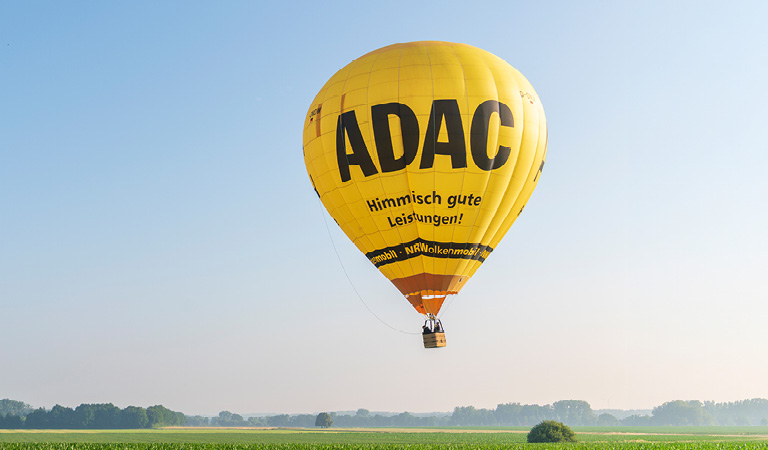 Ein gelber Heissluftballon mit ADAC Logo schwebt über grünen Feldern im Morgenlicht