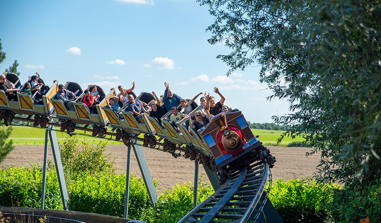 Personen in einer Achterbahnwagen lächeln und heben die Arme inmitten einer grünen Landschaft.