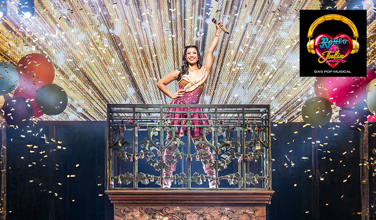 Eine fröhliche Frau in einem bunten Kleid steht singend auf einem Balkon mit Glitzerregen