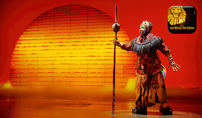 Eine Darstellerin in buntem Kostüm steht ausdrucksstark vor orangeroter Sonne