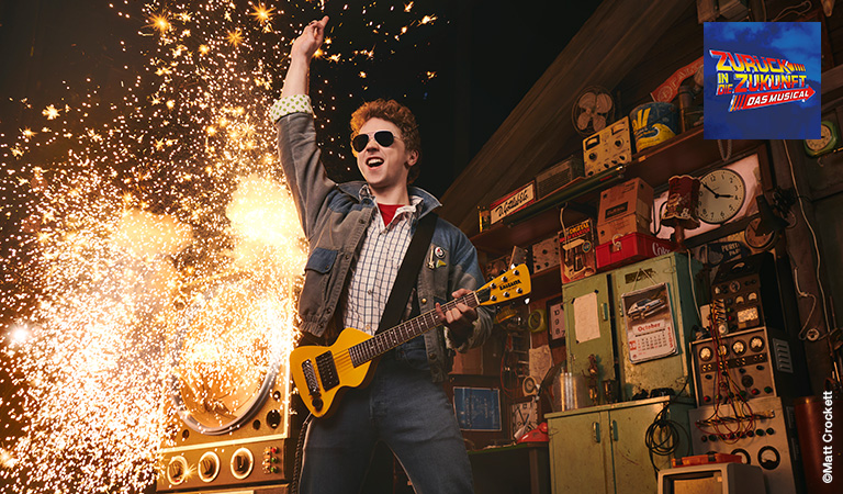 Junger Mann mit roter Lockenfrisur rockt energiegeladen mit gelber Gitarre und Sonnenbrille vor sprühenden Funken