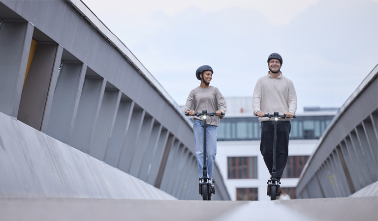 Zwei Personen mit neutralem Ausdruck fahren auf schwarzen E-Scootern über eine graue Brücke