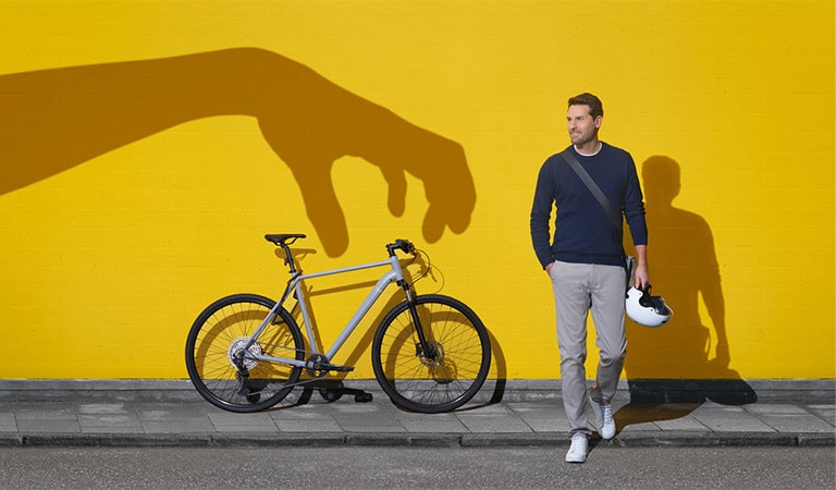 Ein Mann in Freizeitkleidung neben einem Fahrrad vor einer gelben Wand mit großer Hand-Schattenprojektion.