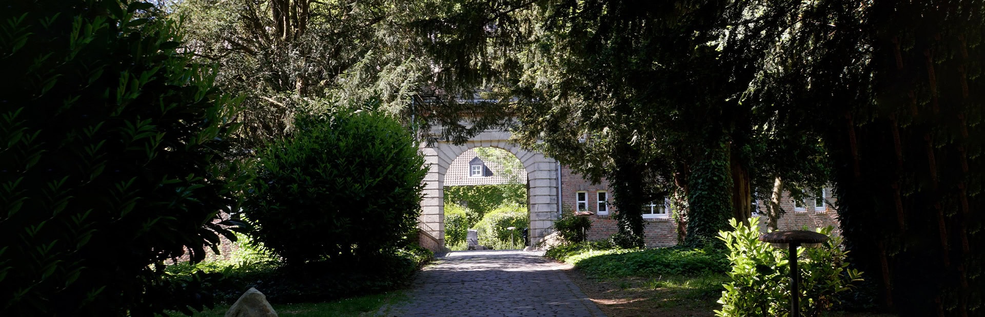 Steinbogen umgeben von üppigem Grün und Bäumen, mit gepflastertem Weg, sonnig und einladend.