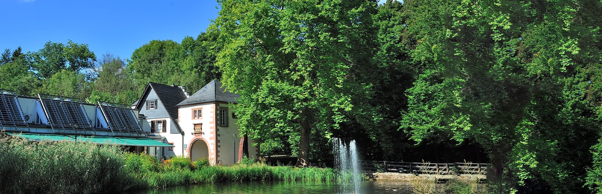 Ein malerisches Haus am Teich mit Springbrunnen, umgeben von grünen Bäumen und blauem Himmel.