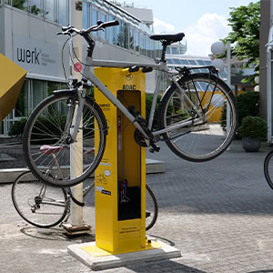 Fahrrad hängt an gelber Reparatursäule vor modernem Gebäude, sonniger Tag.