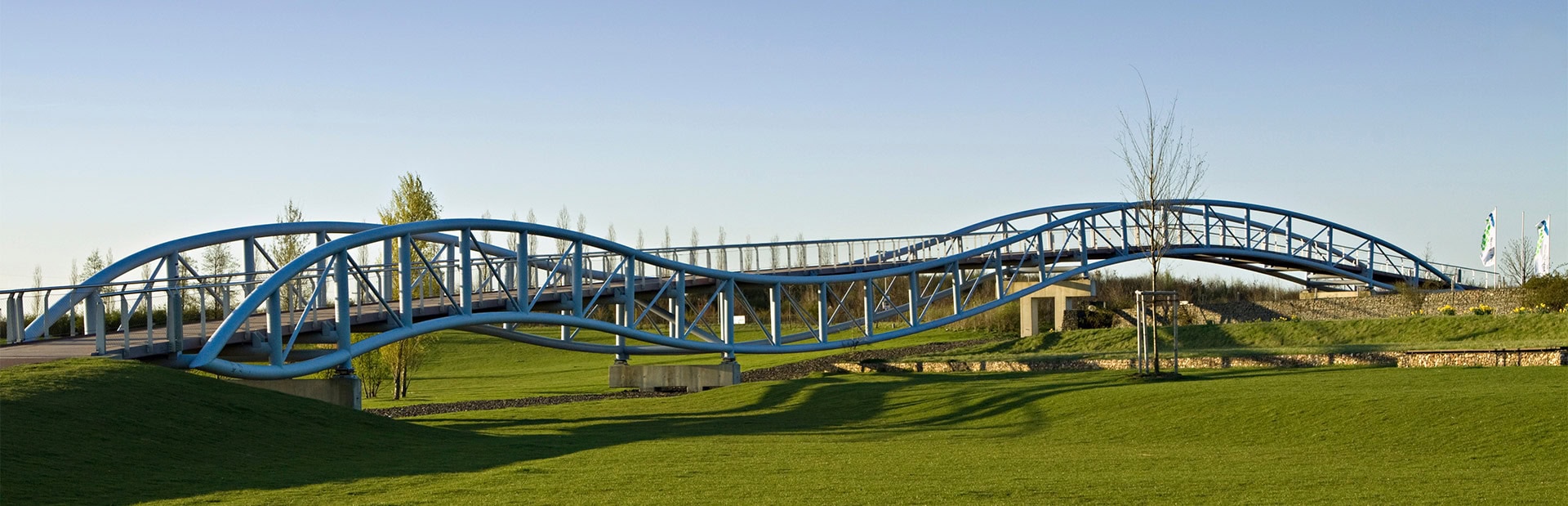 Eine geschwungene, blaue Brücke überspannt grüne Wiesen bei klarem Himmel.