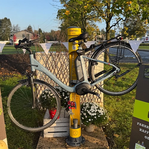 Ein blaues Fahrrad steht an einer gelben Ladestation auf grünem Rasen, umgeben von Blumen.