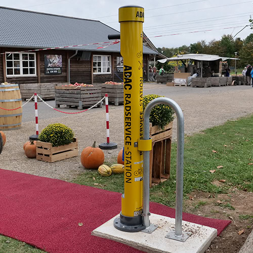 Fahrradservice-Station in Gelb vor einem rustikalen Gebäude mit Kürbissen und Pflanzen.