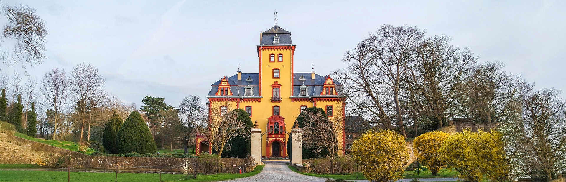 Schloss mit gelben Wänden, roten Details, umgeben von Bäumen und Sträuchern unter bewölktem Himmel.