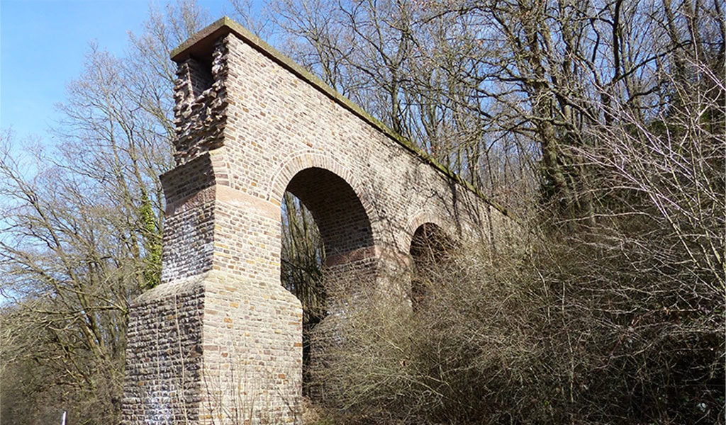 Altes, steinernes Aquädukt in bewaldeter Umgebung, teilweise eingestürzt, bei sonnigem Wetter.