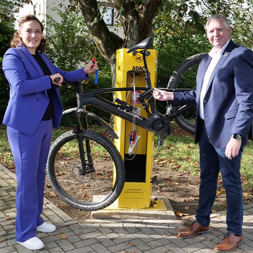 Zwei Personen lächeln in die Kamera, halten ein Fahrrad an einer gelben Reparaturstation.