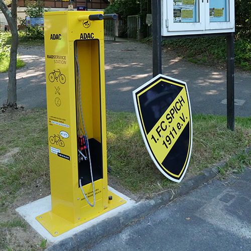 Gelbe ADAC-Fahrradstation, leer. Rechtes Element: Schild "1. FC Spich 1911 e.V." in Gelb-Schwarz.