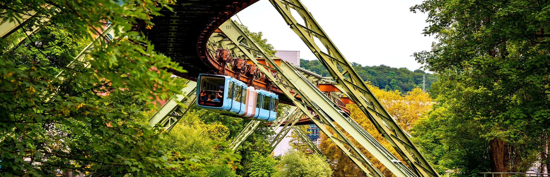Schwebebahn in grüner Umgebung, blaue Wagen, Menschen blicken aus Fenstern, sommerliche Szene.