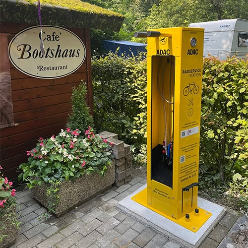 Gelbe ADAC-Fahrradstation vor Café Bootshaus, umgeben von Blumen und Büschen.