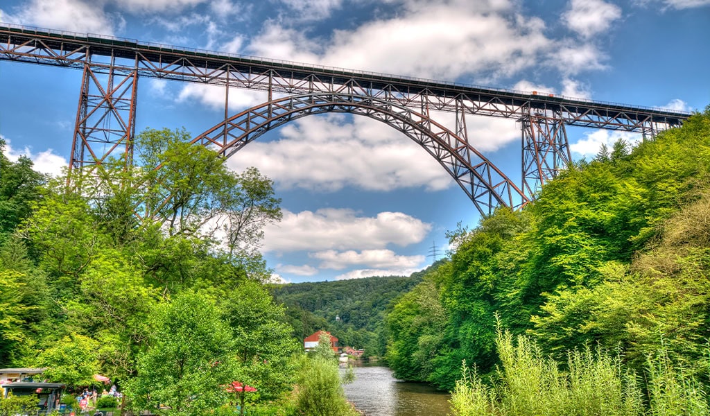 Eine Stahlbrücke überquert ein bewaldetes Tal mit Fluss, klarer Himmel, weiße Wolken.