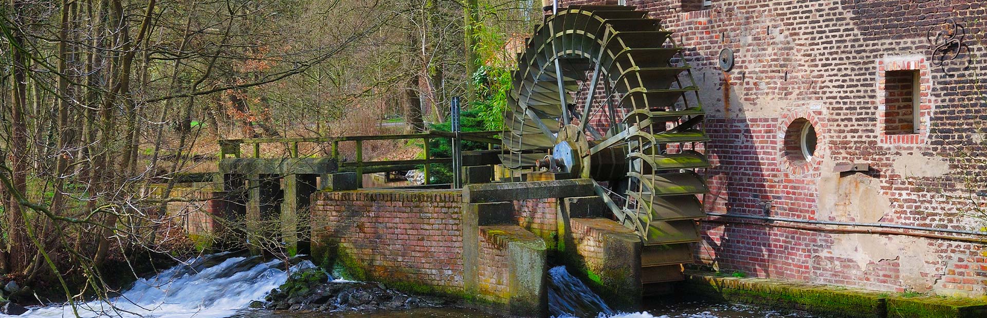 Altes Wasserrad an einem Ziegelbau am Fluss, umgeben von Bäumen in einem ländlichen Gebiet.
