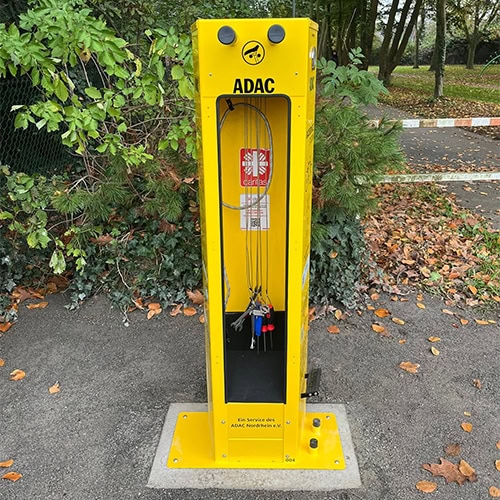 Gelbe ADAC-Ladesäule für E-Bikes mit mehreren Steckdosen und Fahrradinstrumenten, umgeben von Laub.