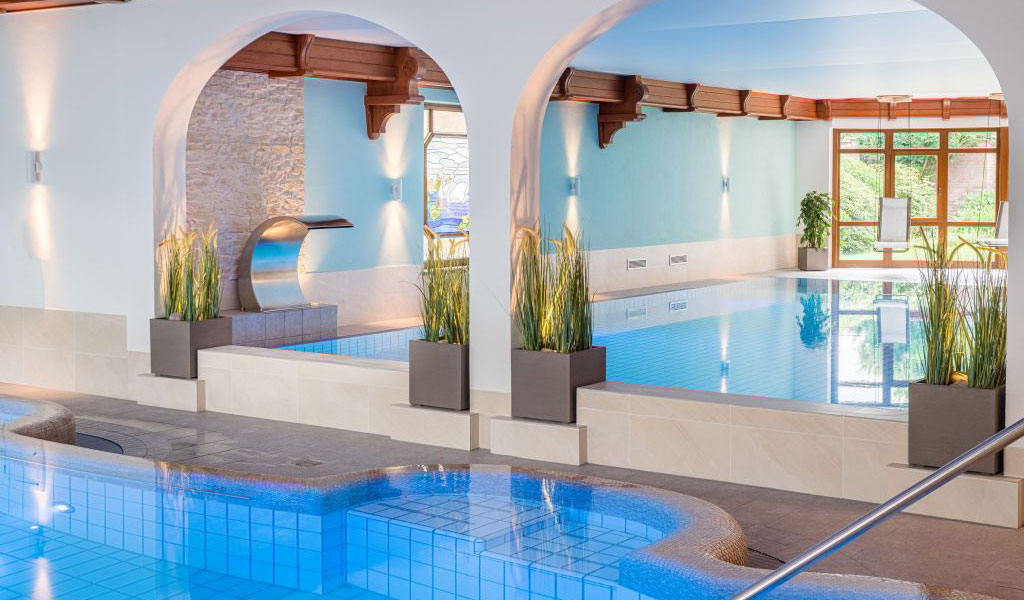 Hallenbad mit blau gefliestem Boden, Lichtbögen, Pflanzen und Wasserfallfunktion, kein Mensch sichtbar.