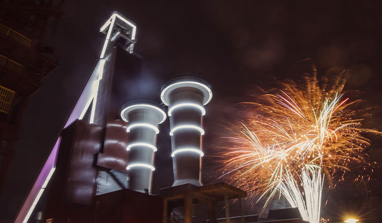 Industrieanlage bei Nacht mit beleuchteten Türmen und buntem Feuerwerk im Hintergrund.
