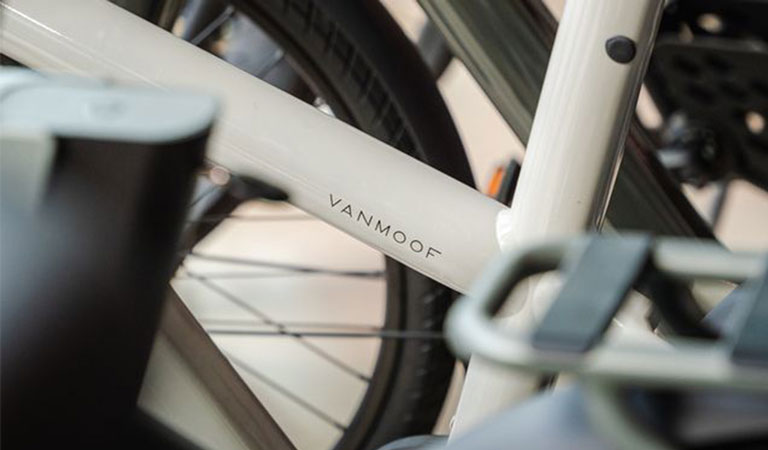 Nahaufnahme eines weißen VanMoof-Fahrradrahmens mit schwarzem "VANMOOF"-Logo, Rad im Hintergrund.