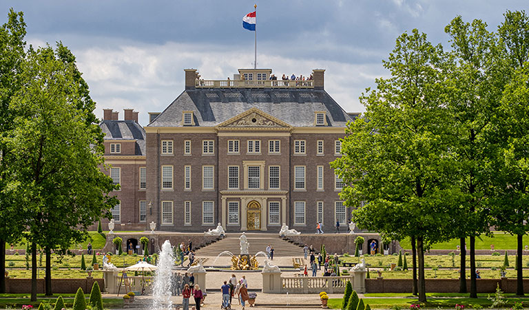 Herrenhaus im Barockstil mit Garten und Springbrunnen; Menschen an der Vorderseite; Flagge oben.