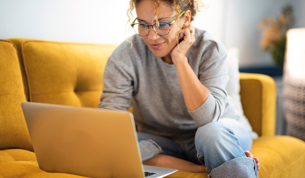 Eine Frau lächelt leicht, sitzt auf einem gelben Sofa und arbeitet am Laptop.
