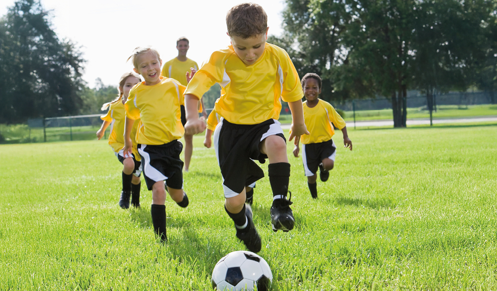 Kinder in gelben Trikots spielen freudig Fußball auf grünem Feld.