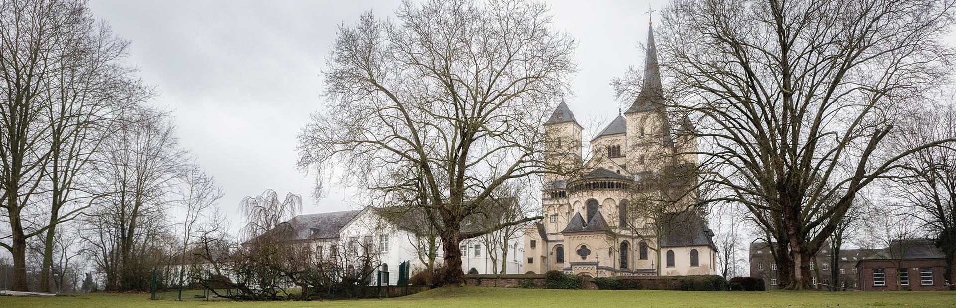 Historische Kirche mit Türmen, umgeben von kahlen Bäumen auf einem grünen Rasen. Wolkiger Himmel.