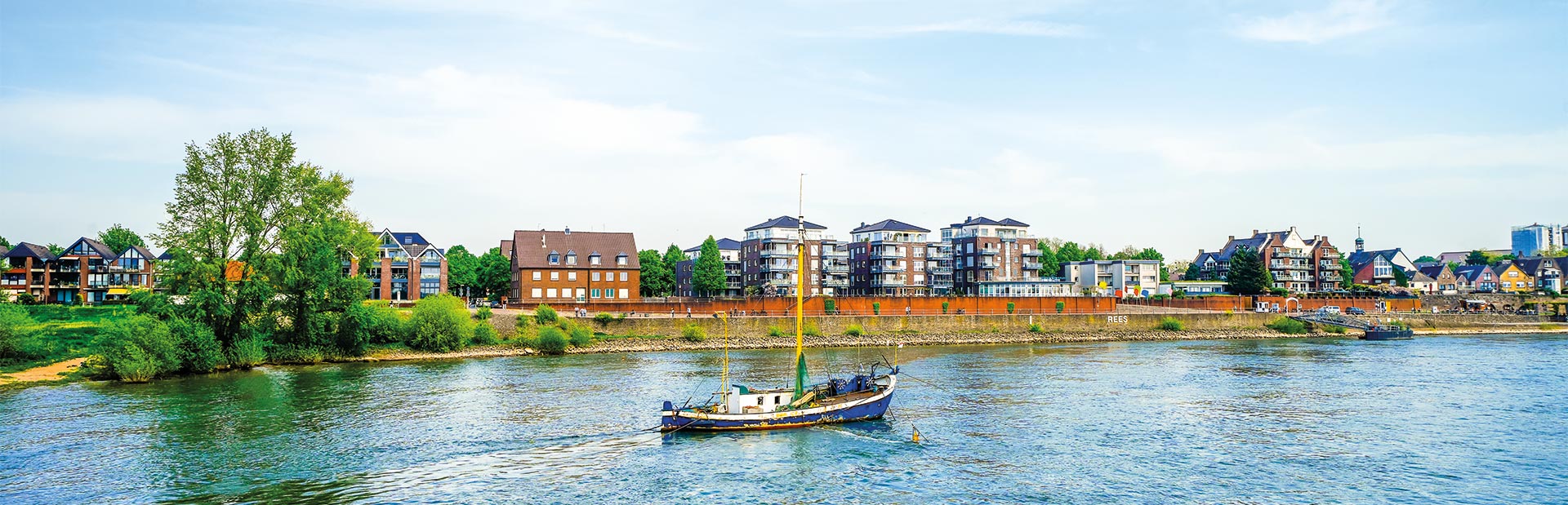Ein Segelboot auf einem Fluss vor einer Stadtlandschaft mit moderner Architektur und klarer Himmel.