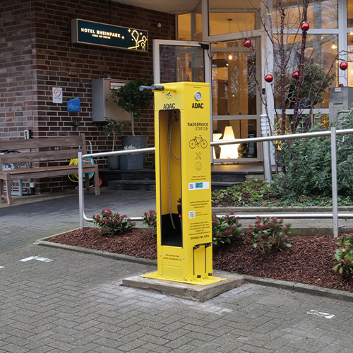 Gelbe Fahrradstation vor einem Hotel, mit roter Weihnachtsdekoration und Pflanzen im Hintergrund.