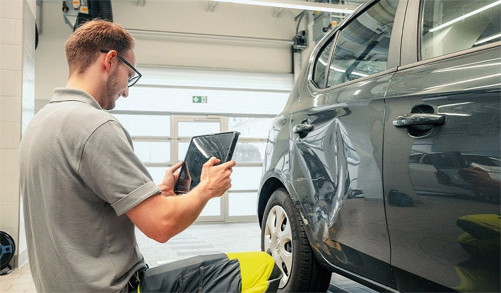 Mann inspiziert Auto mit Tablet in Werkstatt, graues Auto, gelbes Reflexband, konzentrierter Ausdruck.