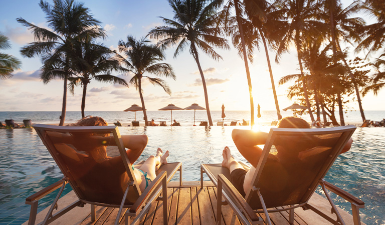 Zwei Personen entspannen am Pool bei Sonnenuntergang mit Palmen und Meer im Hintergrund.