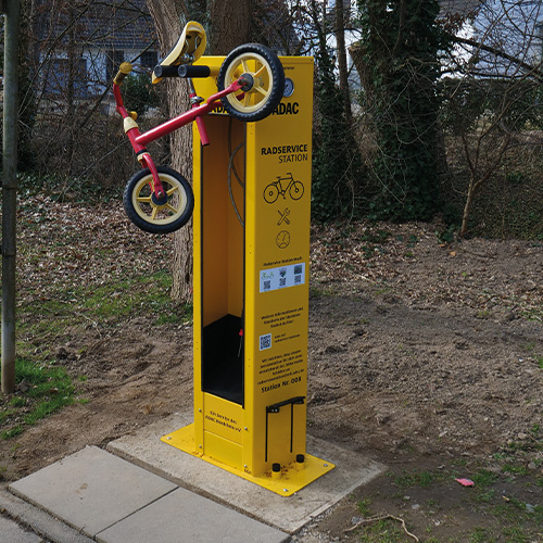 Gelbes Reparatursäule mit aufgehängtem Kinderfahrrad in bunter Umgebung.