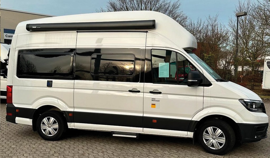 Weißes Wohnmobil mit Hochdach, große Fenster, geparkt auf einem gepflasterten Parkplatz.