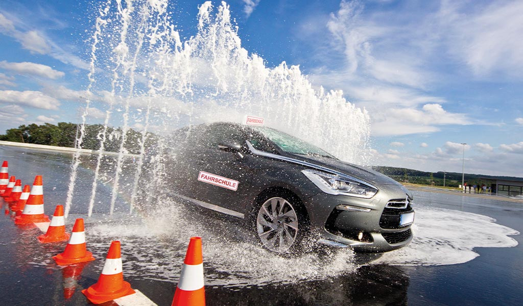 Auto auf nasser Fahrbahn im Schwall von Wasser, umgeben von Warnkegeln, bewölkter Himmel.