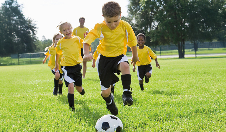Eine Gruppe Kinder in gelben Trikots spielt fröhlich Fußball auf einem grünen Rasenplatz.