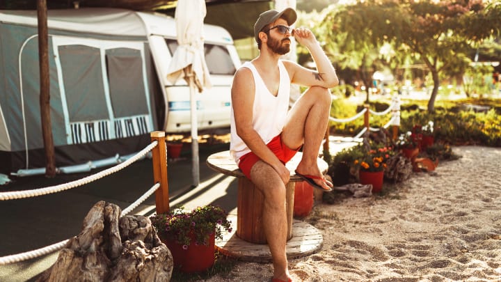 Ein Mann im Freizeitstil, mit Sonnenbrille, sitzt entspannt auf einer Spule neben einem Wohnwagen.