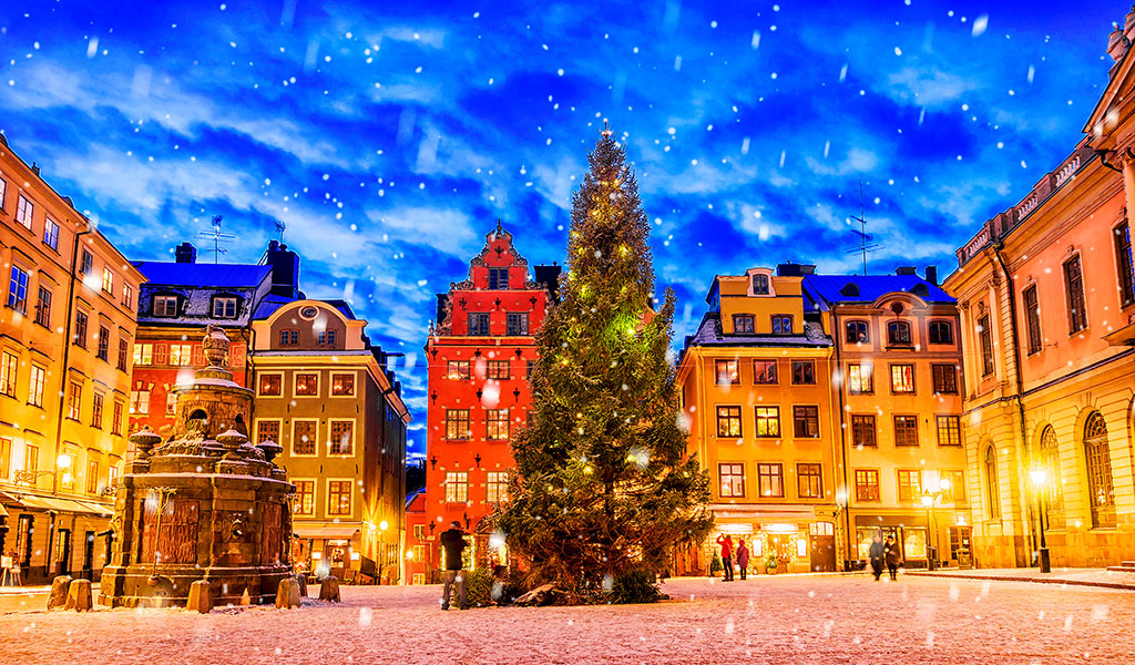 Schneebedeckter Platz mit bunt erleuchteten Gebäuden und Tanne unter nächtlichem Himmel.