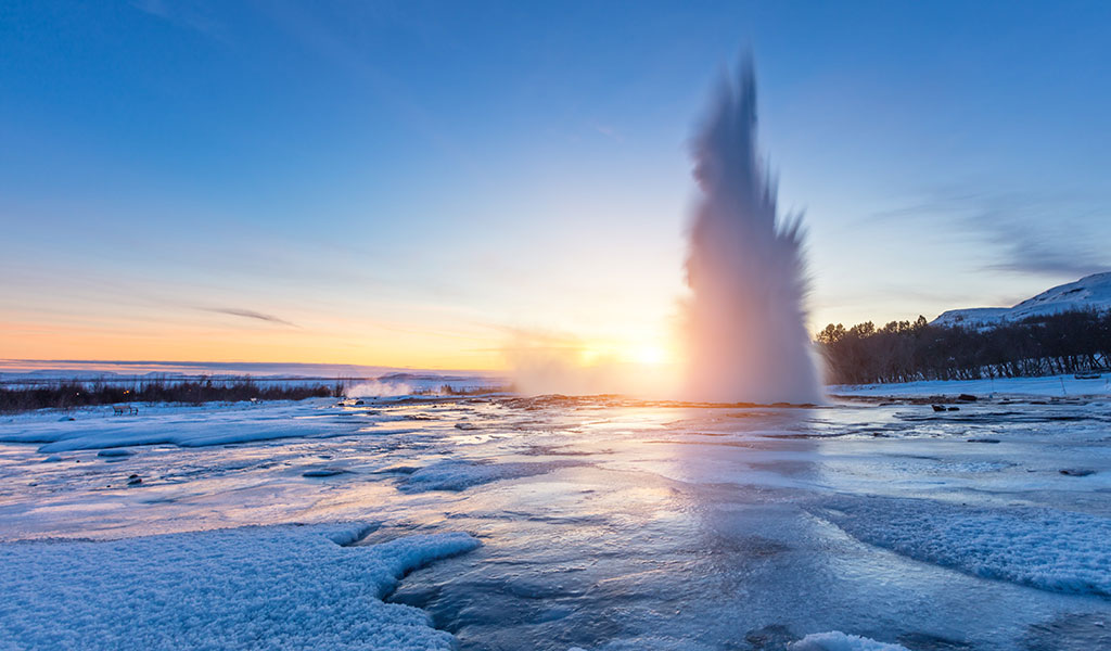 Ein Geysir bricht im Sonnenuntergang aus, umgeben von Schnee und Eis.