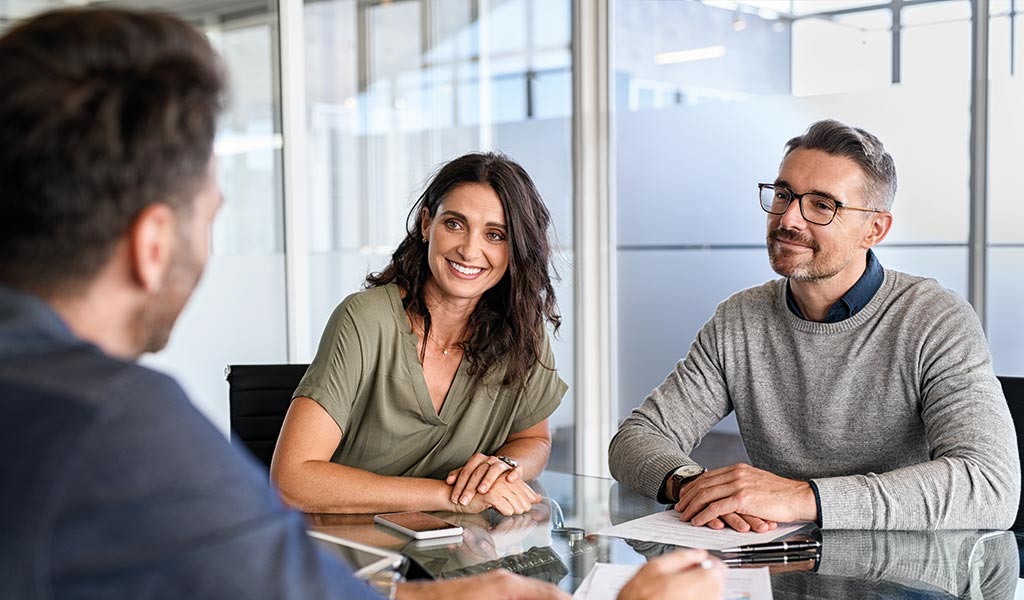 Drei Personen in einem Büro, zwei lächeln freundlich, eine Person sitzt gegenüber.