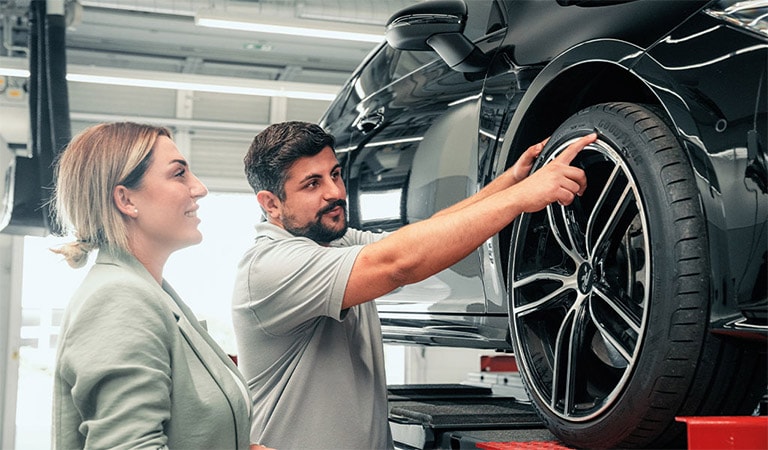 Personen inspizieren ein Auto; Mann zeigt auf Rad, Frau lächelt. Werkstattumgebung, beide fokusiert.