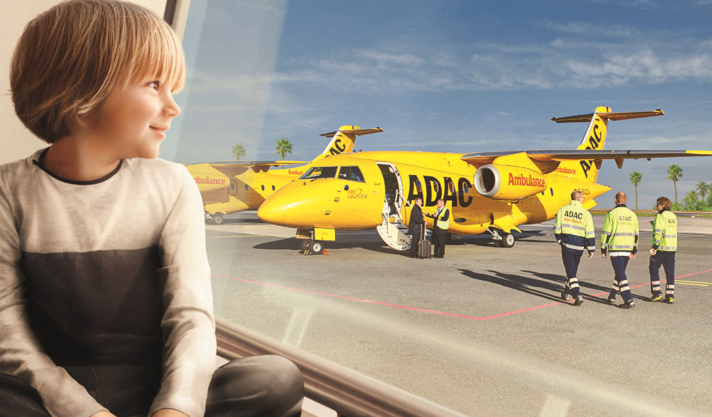 Kind lächelt am Fenster; Blick auf gelbes ADAC-Flugzeug mit drei Besatzungsmitgliedern.