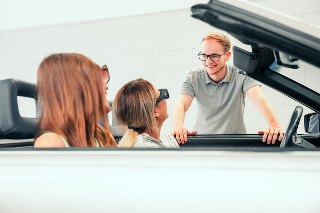 Ein Mann mit Brille lächelt, während er mit drei Frauen in einem weißen Cabrio spricht.