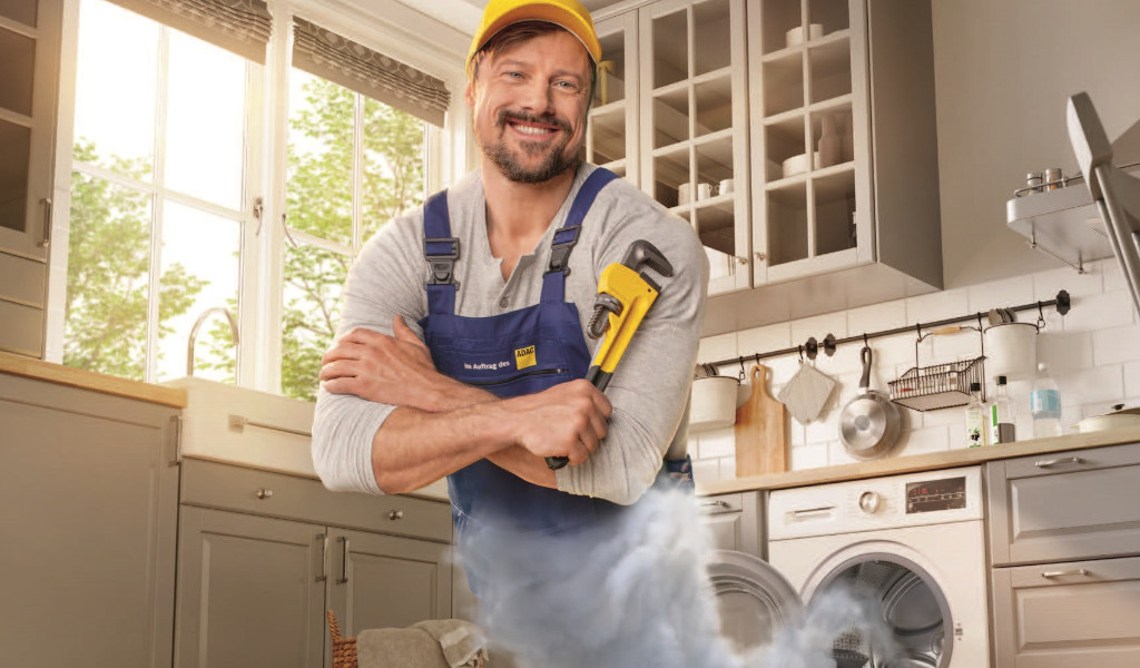 Ein lächelnder Handwerker in gelber Kappe und blauer Latzhose steht in einer rauchenden Küche.