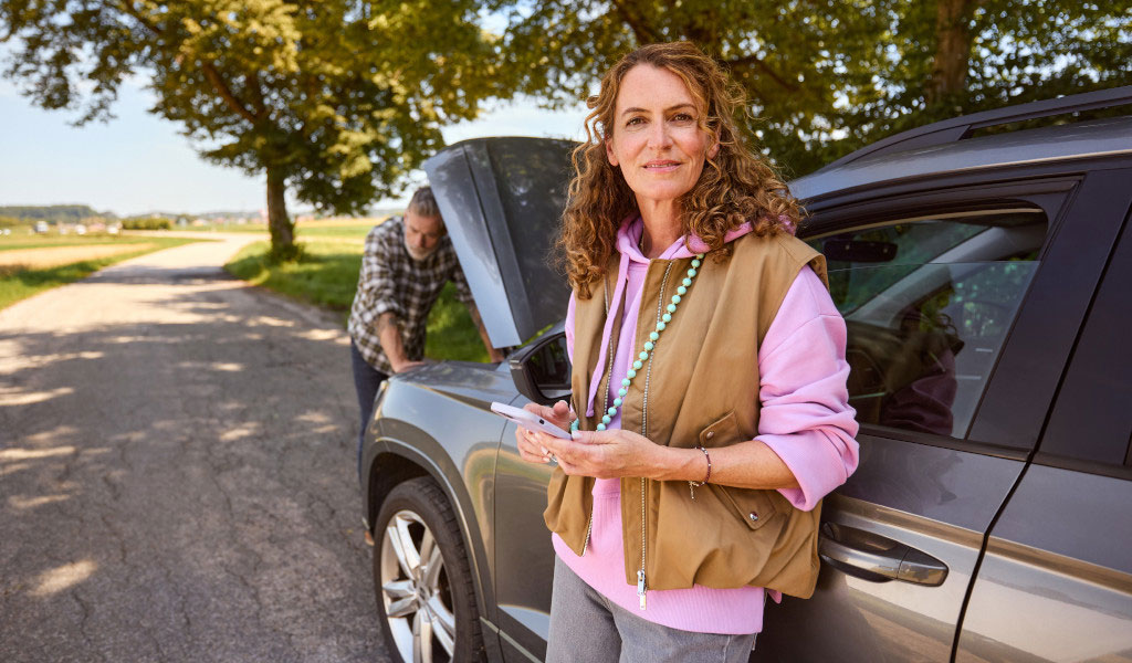 Eine Frau mit lockigem Haar lächelt, während ein Mann am Auto arbeitet.