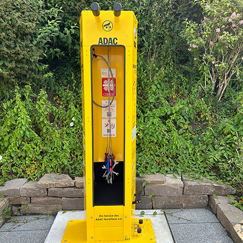 Ein gelbe ADAC-Ladestation mit Schläuchen vor grünen Pflanzen und Steinen